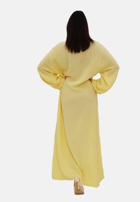 Abito giallo lungo e fluente, realizzato in tessuto testurizzato, con maniche larghe, visto da dietro, abbinato a scarpe con tacco beige.