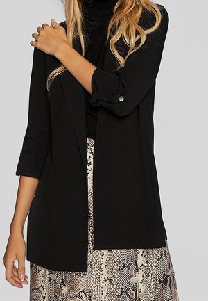 Vrouw draagt een zwarte blazer met opgerolde mouwen over een zwarte top, gecombineerd met een beige en bruine rok met slangenhuidpatroon.