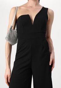Svart jumpsuit med djup urringning och en axelrem. Matchas med en liten, glittrig silverväska. Slät tygtextur.