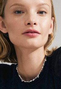 Sidabrinė apyrankė su perlų ir dekoratyvinių akcentų elementais, dėvima aplink kaklą. Paprastas dizainas su pakaitomis tekstūromis ir formomis.