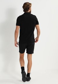 Homme s'éloignant, portant un polo noir, un short noir, des chaussettes noires et des baskets Lacoste noires sur un fond blanc.