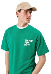 Joven con camiseta verde que dice "FOUND NOT LOST" y gorra beige, mirando hacia su derecha sobre un fondo blanco.