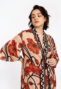 Beige bloemenbadjas met grote rode bloemenprint en zwarte omlijning. Kenmerkend zijn de brede mouwen en een gestructureerde zwarte afwerking langs de halslijn.