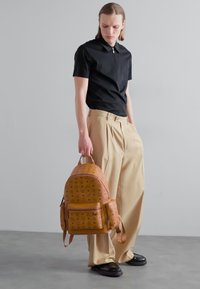Zaino in pelle marrone con motivo a rilievo, forma arrotondata e tasca frontale; pantaloni beige a vita alta e polo nera.