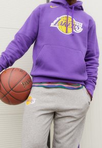 Fioletowa bluza z kapturem z logo Los Angeles Lakers, szare spodnie dresowe z kolorowymi paskami w pasie, trzymająca piłkę do koszykówki.