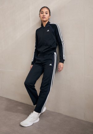 Tuta sportiva nera con strisce bianche, composta da una giacca con zip e pantaloni con vita elasticizzata, abbinata a scarpe da ginnastica bianche.