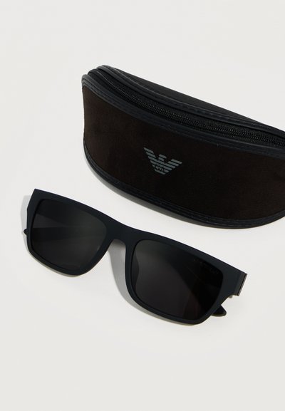 Μαύρα ορθογώνια γυαλιά ηλίου δίπλα σε μαύρη σουέντ θήκη με φερμουάρ και ασημένιο λογότυπο αετού σε λευκό φόντο.