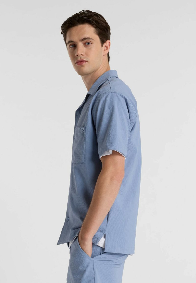 Chemise bleu clair à manches courtes en tissu texturé avec une poche poitrine et une fermeture par boutons. Associée à un pantalon bleu assorti.