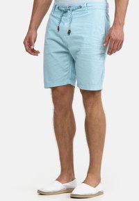 Homme portant un short bleu clair avec cordon de serrage et des chaussures blanches sans lacets, debout devant un fond blanc uni.