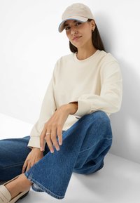 Beige sweatshirt met lange, iets opbollende mouwen. Beige pet met blauwe accenten. Losse blauwe denim jeans. Stijlvol, strak ontwerp.
