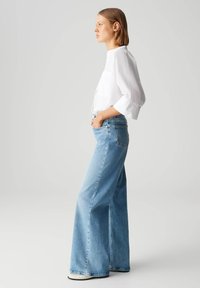 Chemisier blanc à boutons avec des manches trois-quarts, associé à un jean en denim bleu clair à jambes larges, doté de poches avant et d'une coupe décontractée.