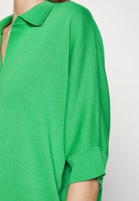 Polo shirt verde lavorato a maglia con colletto, maniche corte e polsini a costine. Il tessuto ha una consistenza liscia e un colore uniforme.