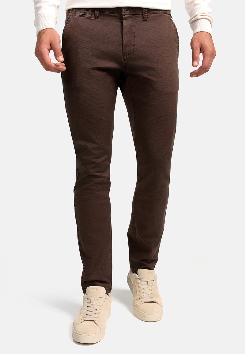 Brown slim-fit chinos made of smooth fabric, featuring a flat front and subtle texture. Paired with light beige sneakers.