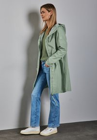 Veste imperméable à capuche vert clair, avec fermeture éclair à l'avant, associée à un pull vert et un jean bleu. Baskets blanches avec des semelles texturées.