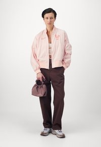 Pink bomber jacket with a ribbed collar, brown wide-leg trousers, and a small purple gathered bag; white sneakers with accents.