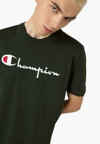 T-shirt in cotone verde scuro con testo ricamato bianco "Champion" e logo sull\'orlo della manica sinistra. Maniche corte, scollo rotondo.