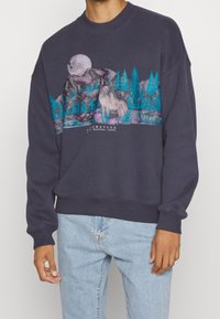 Granatowy sweter z okrągłym dekoltem, z nadrukiem graficznym przedstawiającym wilka, góry i drzewa w żywych kolorach. Miękka tekstura z ściągaczami przy mankietach.