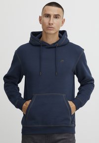 Mornarsko moder kapuco s sprednjim žepom, kontrastnim šivom in kapuco na vrvico. Značilna je majhna oznaka na prsih.