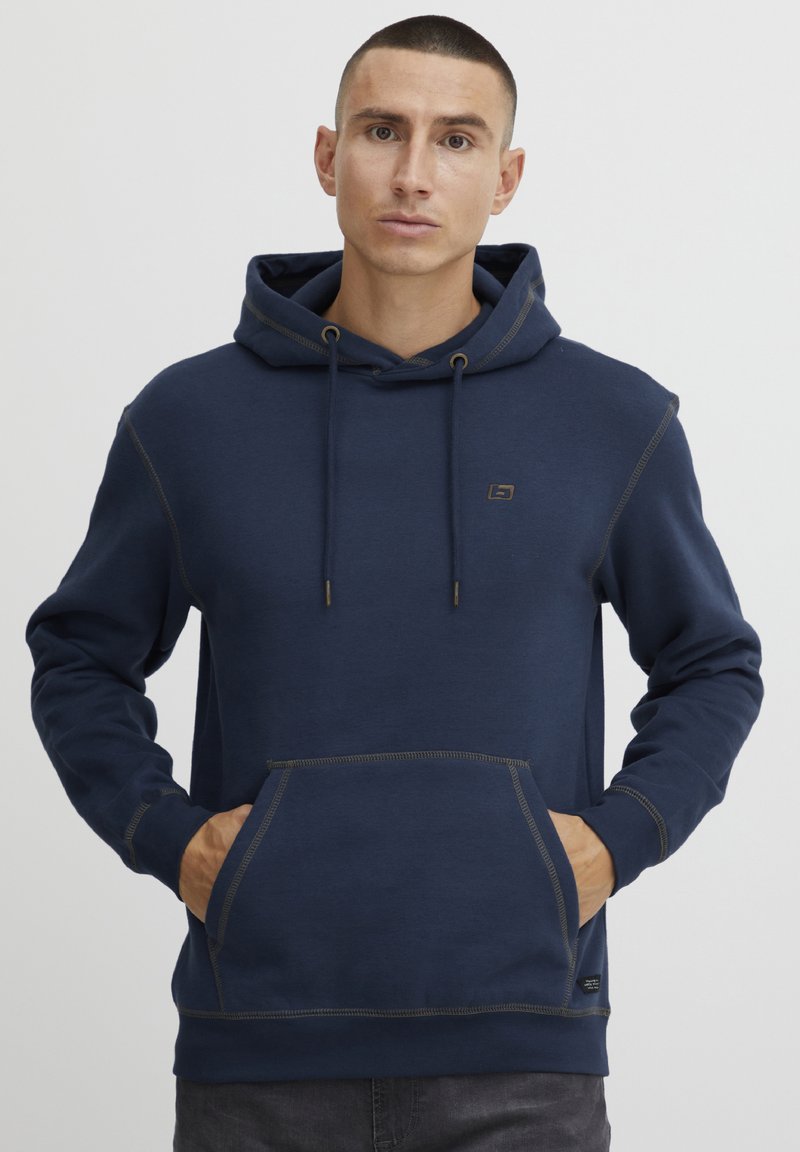 Mornarsko moder kapuco s sprednjim žepom, kontrastnim šivom in kapuco na vrvico. Značilna je majhna oznaka na prsih.