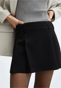 Falda corta negra con pliegues y una pretina estructurada. La tela parece suave con un acabado mate, combinada con una blusa gris ajustada.