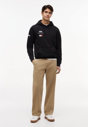 Young man standing with hands partially in pockets, wearing a black hoodie with logos, beige pants, and white sneakers on a white background.