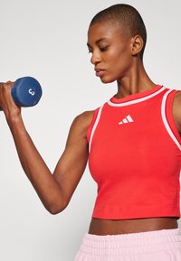Piros sportos crop top fehér díszítéssel, kerek nyakkivágással és testhezálló fazonnal. A modellt az egyik kezében egy kék súlyzót tart.