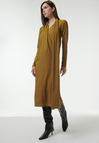 Vestido maxi de color mostaza amarillento con mangas largas y un escote drapeado, combinado con botas de tacón negro hasta los muslos. Textura suave, silueta ajustada.