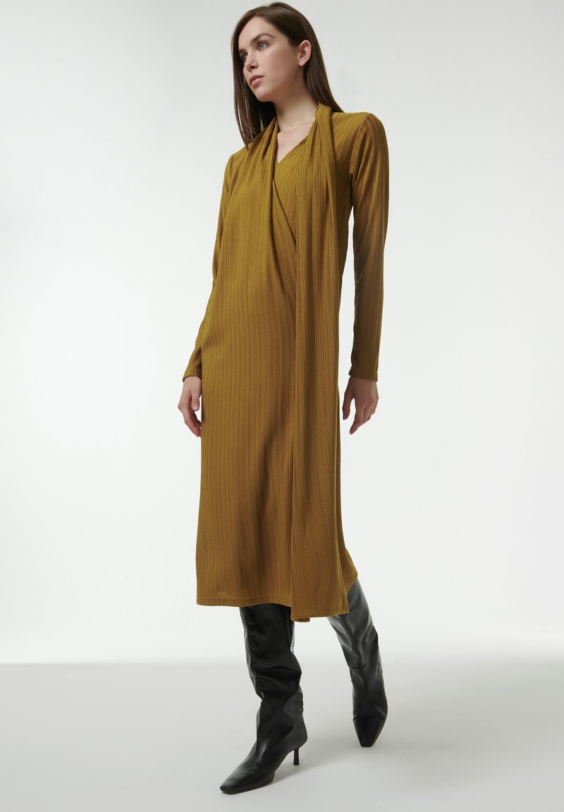 Vestido maxi de color mostaza amarillento con mangas largas y un escote drapeado, combinado con botas de tacón negro hasta los muslos. Textura suave, silueta ajustada.