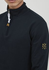 Mornarska zip-up majica od mekog materijala s visokim kragnom i bijelom šniranjem. Ima žuti izvezeni logo na lijevoj rukavi.