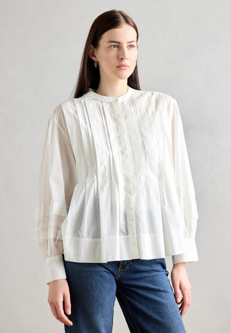 White, pleated blouse with a high collar and button closure, featuring sheer panels and long sleeves. Paired with blue jeans.