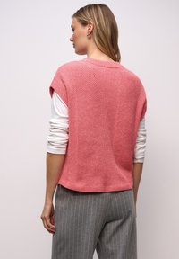 Růžový pletený top bez rukávů s texturovaným vzorem, nošený přes bílou košili s dlouhým rukávem, doplněný šedými kalhotami s proužky.