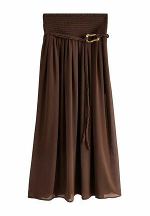 Frau mit langem braunem Haar, die ein weißes ärmelloses Oberteil und einen fließenden braunen Maxirock trägt, seitlich vor einem neutralen Hintergrund stehend.