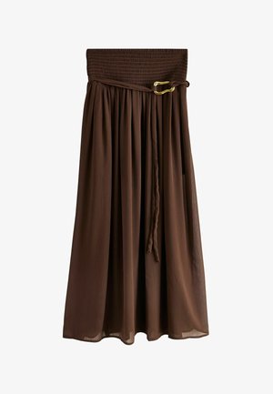 Kvinde med langt brunt hår iført en hvid, ærmeløs top og en flydende brun maxinederdel, stående sidelæns mod en neutral baggrund.