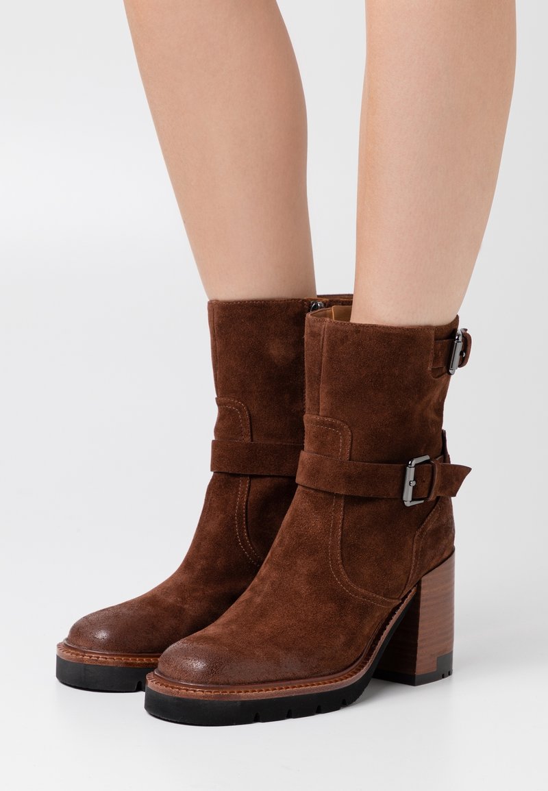 Bottes mi-mollet en daim marron avec talons blocs, brides frontales et boucles argentées portées sur jambes nues sur fond uni.
