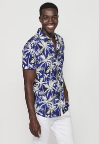 Camicia a maniche corte con bottoni, di colore blu con motivo di palme bianche. Realizzata in tessuto leggero, con colletto classico e bottoni frontali.