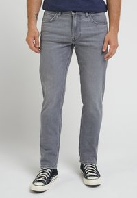 Man wearing slim-fit grey jeans and black sneakers with white laces, standing against a plain white background.