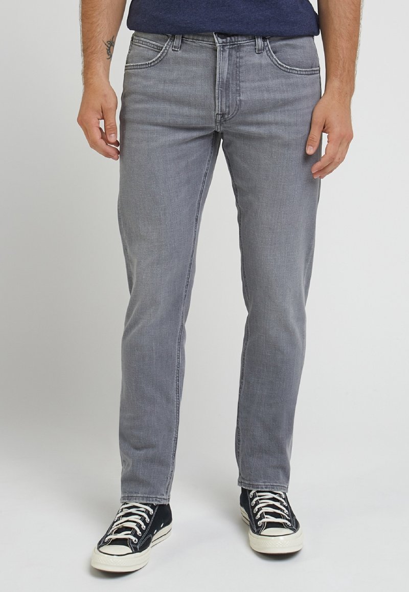 Man wearing slim-fit grey jeans and black sneakers with white laces, standing against a plain white background.