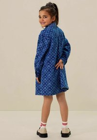 Robe en denim bleu avec motif floral, col, manches longues et coupe décontractée. Portée avec des chaussettes blanches et des chaussures noires, le modèle a les cheveux tirés en arrière.