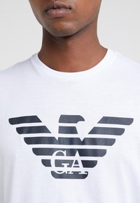 T-shirt blanc avec un design graphique noir représentant des ailes d'aigle stylisées et les lettres "GA." Texture douce, encolure ronde.