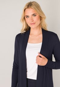Marineblauwe cardigan gemaakt van een zachte, lichtgewicht stof, met een open voorkant en lange mouwen, gecombineerd met een wit topje. Eenvoudige gouden ketting.