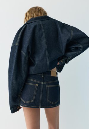 Femme de dos portant une veste en jean oversize foncée et une mini-jupe taille haute assortie avec des surpiqûres jaunes sur un fond uni.