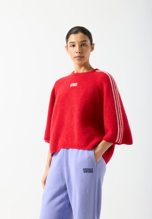 Frau trägt einen roten Kurzarmpullover und lavendelfarbene Hose, beide mit "American Vintage" Logos, steht mit einer Hand in der Tasche.