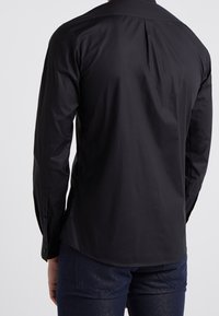 Camicia nera a maniche lunghe con una trama liscia, polsini con bottoni e un dettaglio a piega sul retro. Vestibilità sartoriale con orlo curvo sul fondo.