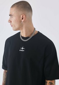 Camiseta de algodón negra de manga corta, con un logo bordado en blanco en el pecho. El modelo lleva puesta una cadena de plata.