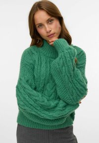 Frau mit hellbraunem Haar, die einen grünen, grob gestrickten Pullover und graue Hosen trägt, mit dem Kinn auf der Hand ruhend vor einem schlichten Hintergrund.
