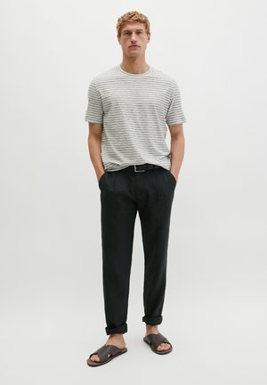 Jonge man staat met zijn handen in zijn zakken, draagt een grijs-wit gestreept T-shirt, zwarte opgerolde broek en donkere slippers.