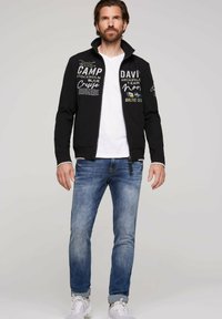 Sort zip-up jakke med hvide og farvede tekstgrafikker, suppleret med en hvid T-shirt og blå jeans, med en afslappet pasform og manchetter på ærmerne.