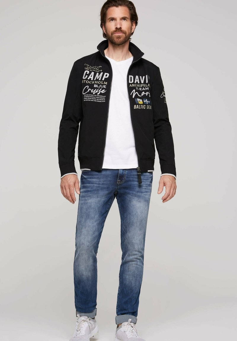 Sort zip-up jakke med hvide og farvede tekstgrafikker, suppleret med en hvid T-shirt og blå jeans, med en afslappet pasform og manchetter på ærmerne.