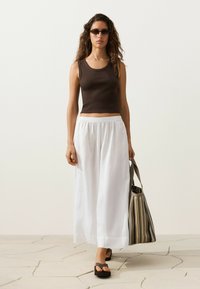 Femme aux longs cheveux bouclés portant des lunettes de soleil foncées, un haut marron sans manches, un pantalon blanc à jambes larges, des sandales noires, et tenant un sac fourre-tout rayé.