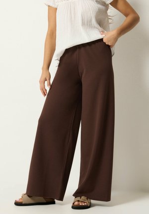 Donna con pantaloni marroni a gamba larga, camicetta bianca con texture e sandali beige con punta aperta, in piedi davanti a uno sfondo semplice.
